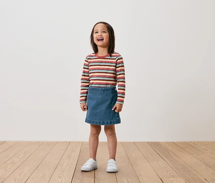 Une fille se tient sur un plancher en bois et porte un t-shirt rayé à manches longues, une jupe en jean et des baskets blanches.
