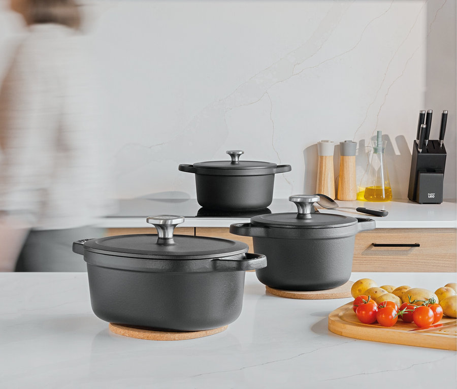 Trois casseroles noires avec couvercles sur un comptoir de cuisine, à côté d'une planche à découper avec des tomates et des pommes de terre.