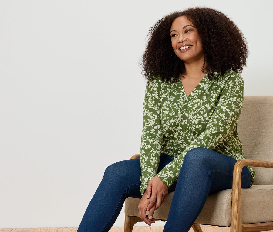Une femme est assise dans un fauteuil. Elle porte une blouse verte à motifs floraux et un jean.