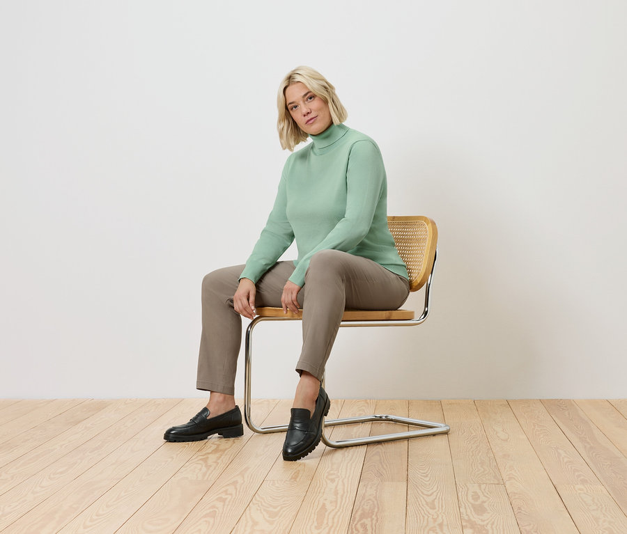 Une femme est assise sur une chaise. Elle porte un pull à col roulé vert, un pantalon marron et des chaussures noires.