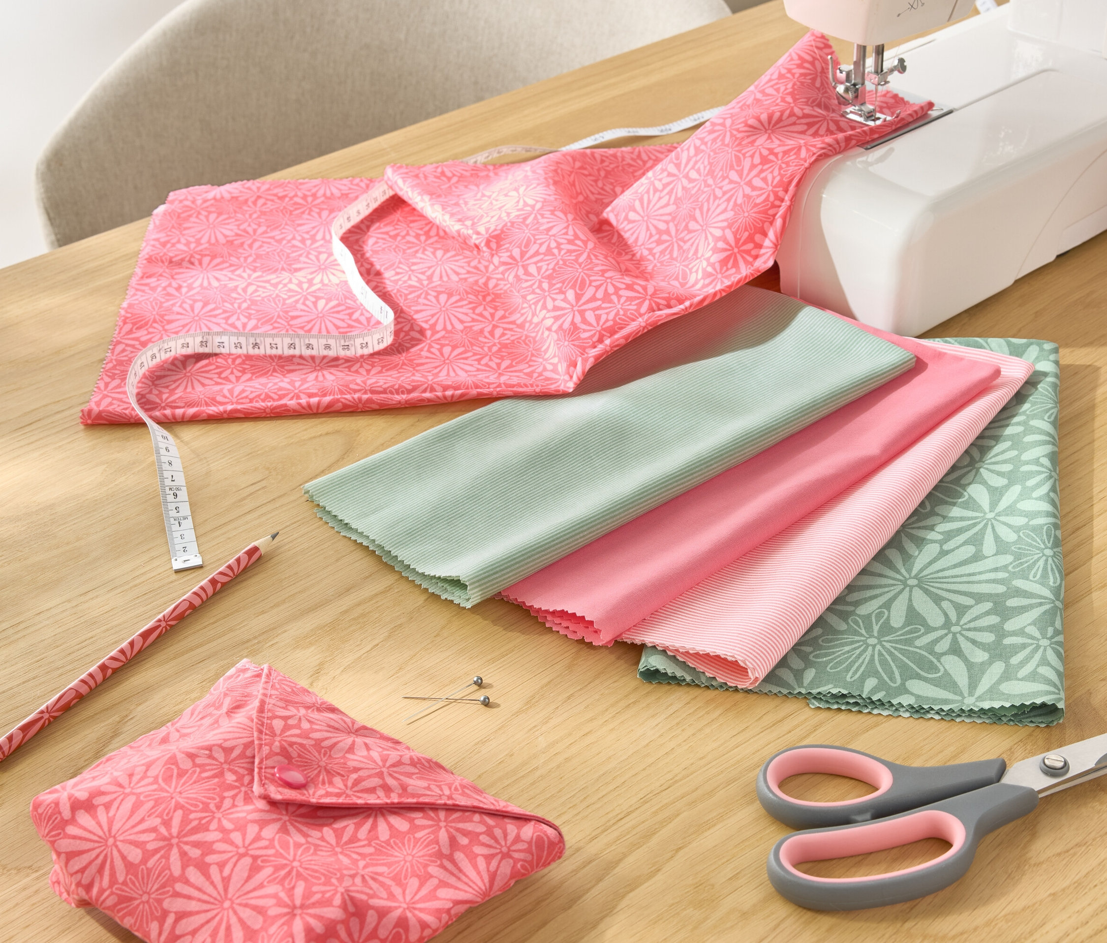 Sur une table sont disposés un ensemble de tissus 5 pièces, une machine à coudre, des ciseaux, un crayon et un mètre ruban.