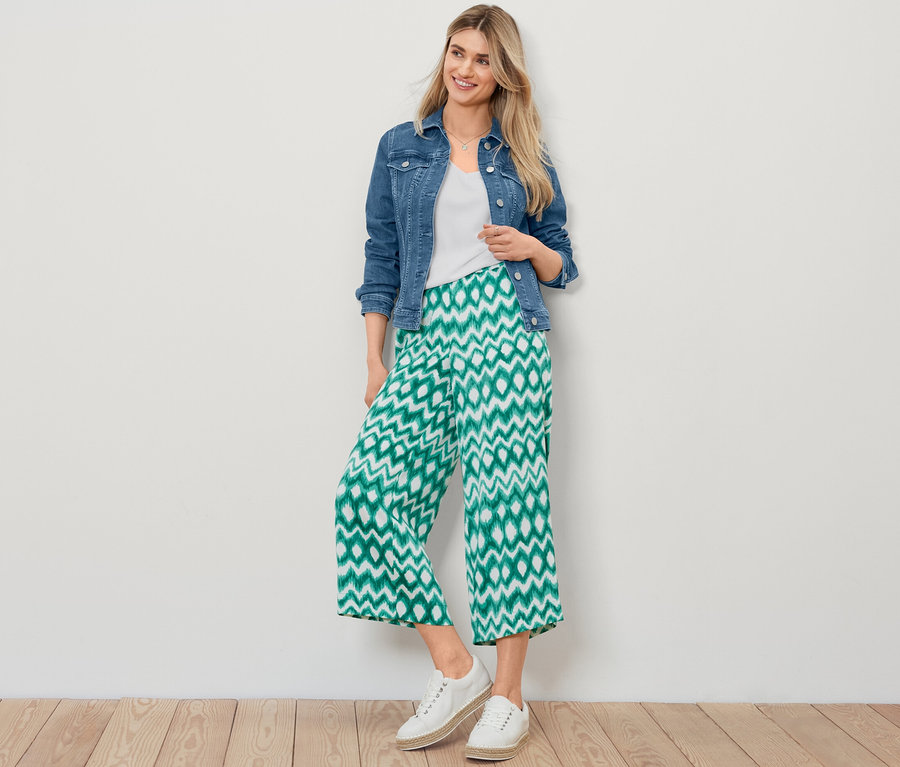 Une femme blonde pose en veste en jean, blouse structurée blanche et pantalon en satin imprimé.