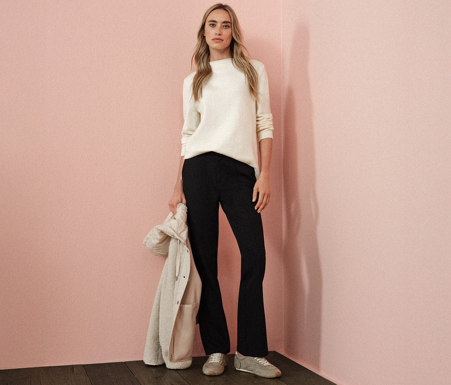 Une femme porte un pull en tricot beige à bord roulé, un pantalon noir et tient une veste.