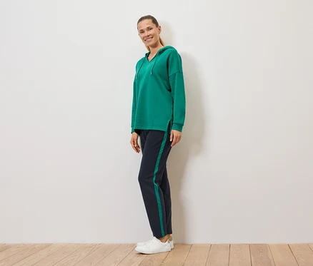 Une femme se tient de profil, portant un sweat à capuche vert et un pantalon de jogging bleu marine avec des bandes latérales vertes.