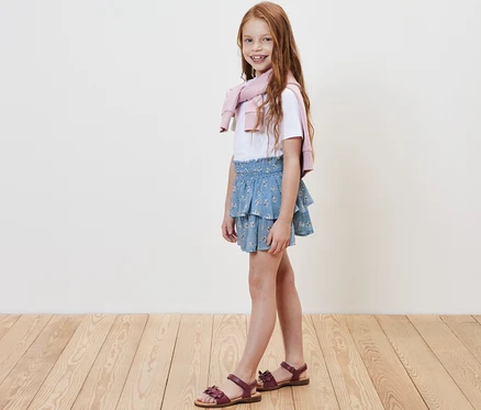 Fillette souriante posant en jupe étagée pour enfant, t-shirt blanc et pull rose jeté sur les épaules.