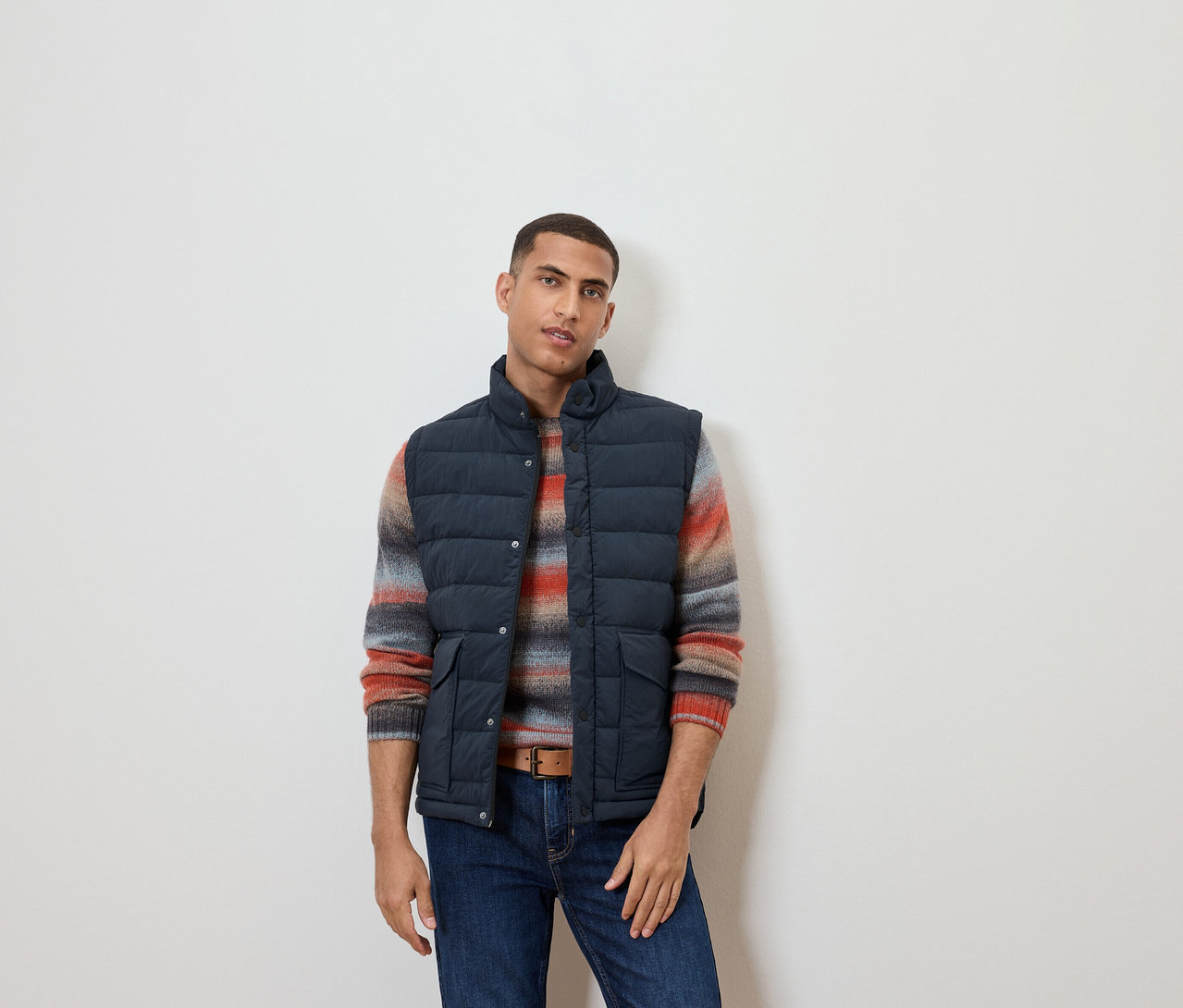 Un homme portant un gilet matelassé sans manches, un pull rayé et un jean.