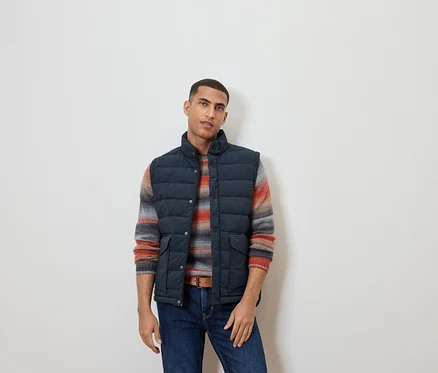 Un homme portant un gilet matelassé sans manches, un pull rayé et un jean.