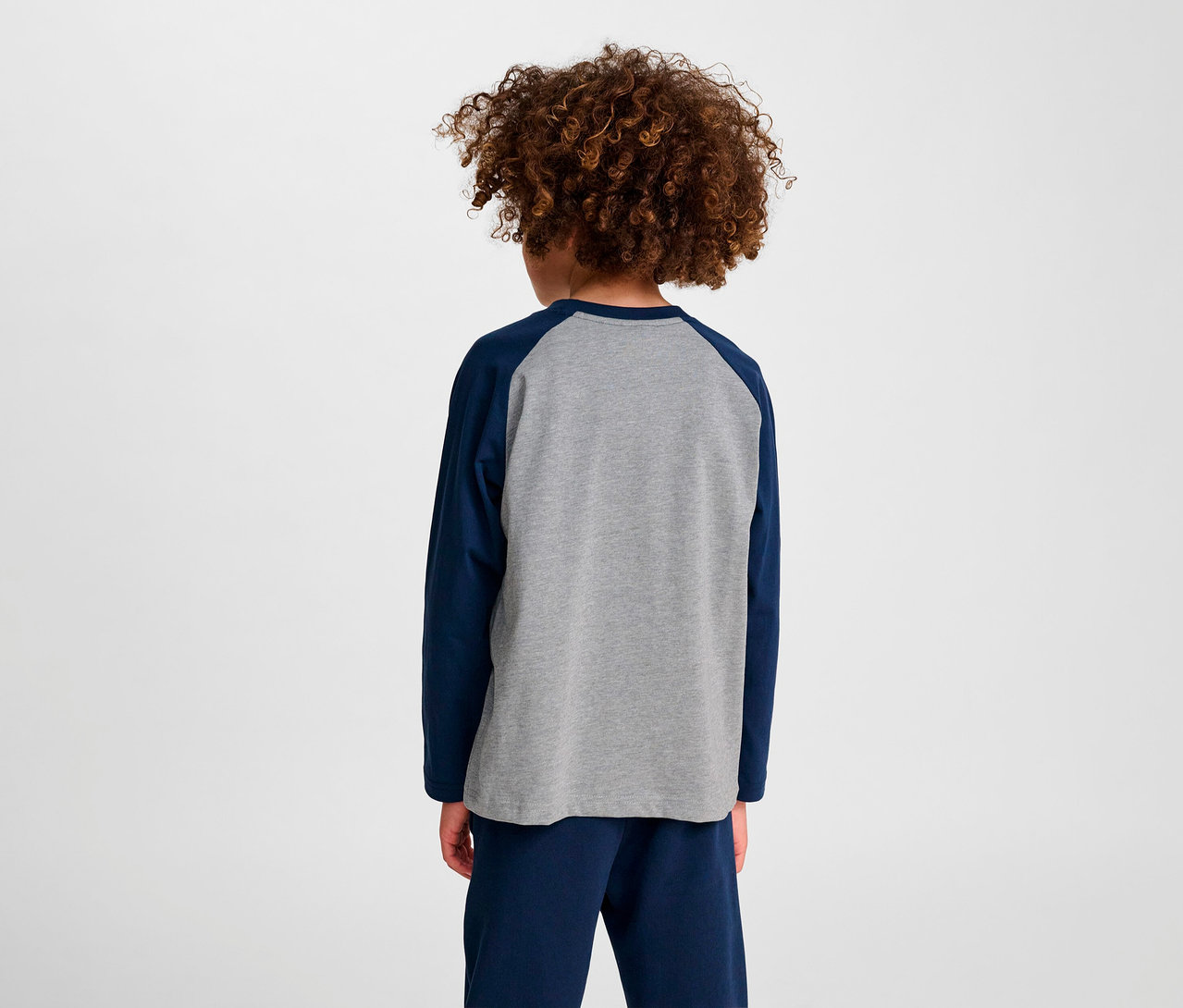 Un garçon aux cheveux bouclés, vu de dos, porte un T-shirt ample à manches raglan HUMMEL HMLJR BEE bleu.