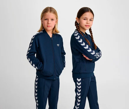 Deux filles posent en studio en survêtements bleus Hummel. Veste à fermeture éclair HUMMEL HMLJR Archive Reg Poly, bleue