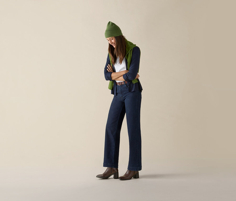 Une femme pose, les bras croisés, portant un pull en maille fine vert clair, un jean et des bottes marron.