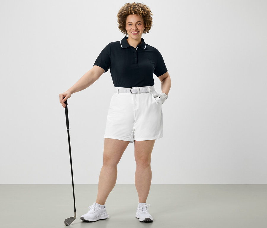 Femme tenant un club de golf, portant un polo technique bleu foncé et un short technique en softshell blanc.