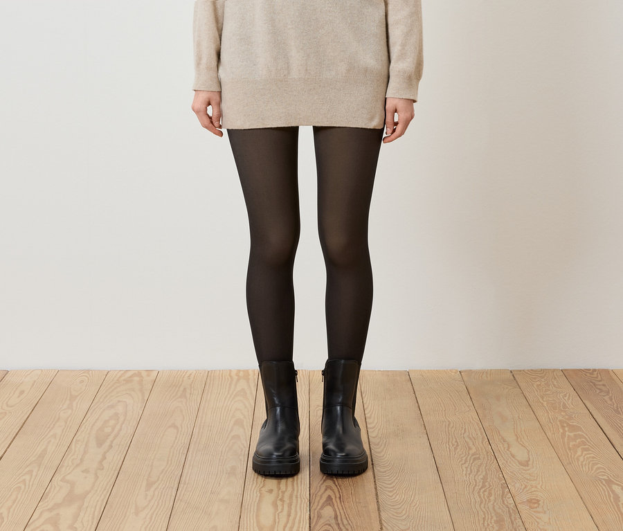 Une femme porte un pull beige, des collants noirs et des bottes noires.