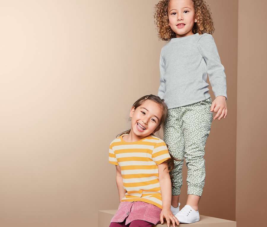 Deux filles posent sur un bloc. L'une porte un 2 t-shirts en matière côtelée gris et un pantalon vert. L'autre porte un t-shirt rayé jaune et une jupe bordeaux.