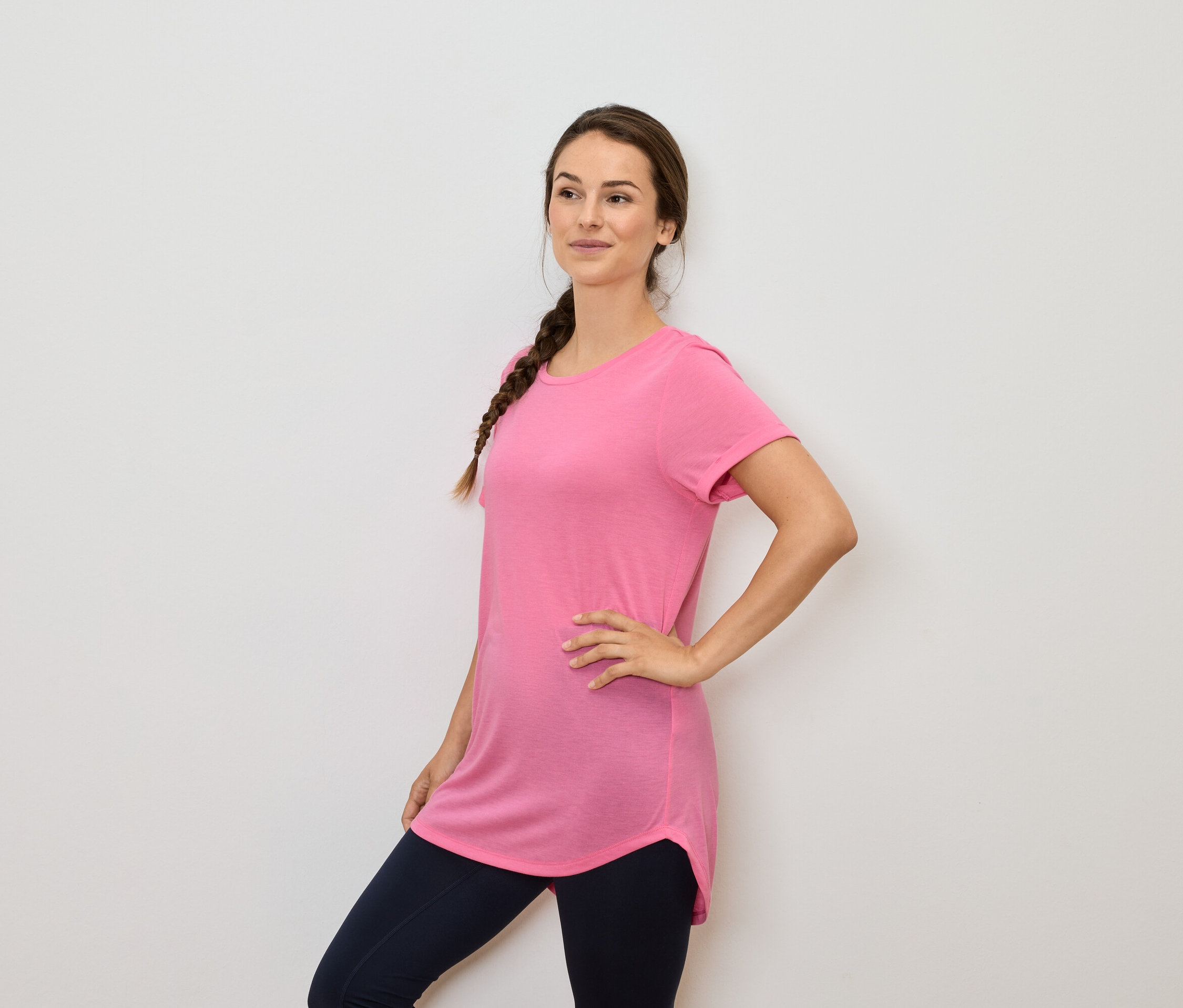 Femme aux cheveux bruns tressés posant dans un t-shirt rose et un legging bleu marine.