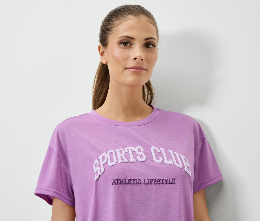 Femme portant un t-shirt lilas avec l'inscription SPORTS CLUB ATHLETIC LIFESTYLE.