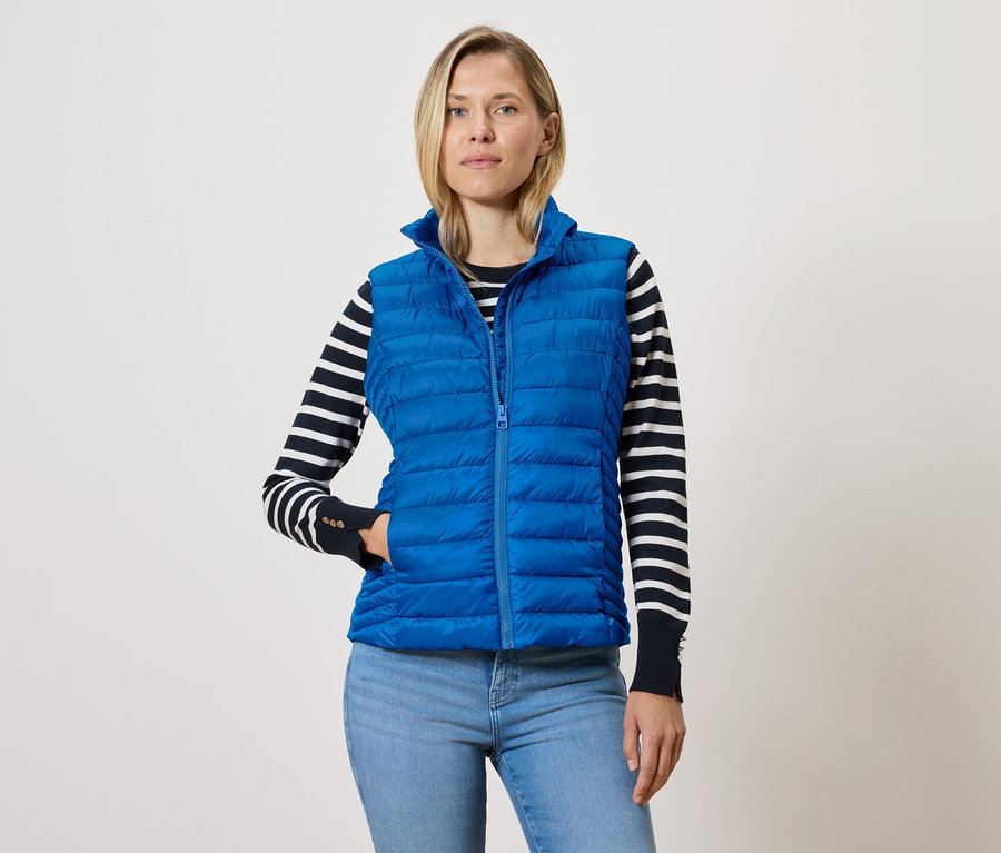 Une femme portant une veste sans manches matelassée bleue avec fermeture éclair, un pull rayé noir et blanc et un jean bleu.