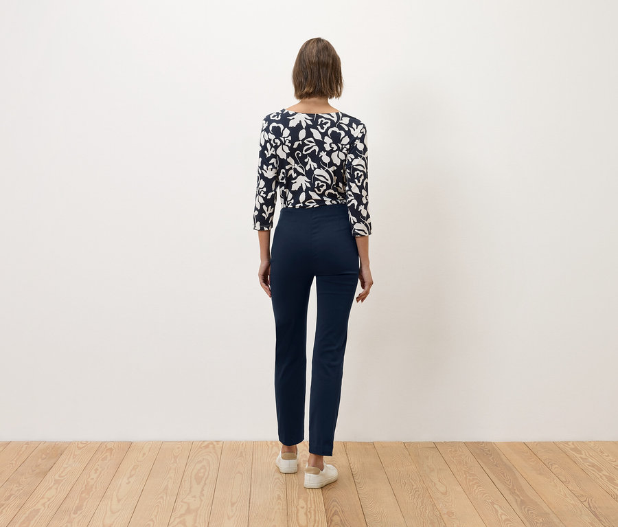 Une femme aux cheveux bruns se tient dos à la caméra, portant un pantalon bleu marine et un haut bleu et blanc à motifs floraux.
