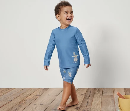 Un jeune garçon souriant, vêtu d'une combinaison bleue pour enfants avec protection UV 80 et imprimé kangourou, marche sur un plancher en bois. À droite, un seau jaune contient des jouets dans le sable.
