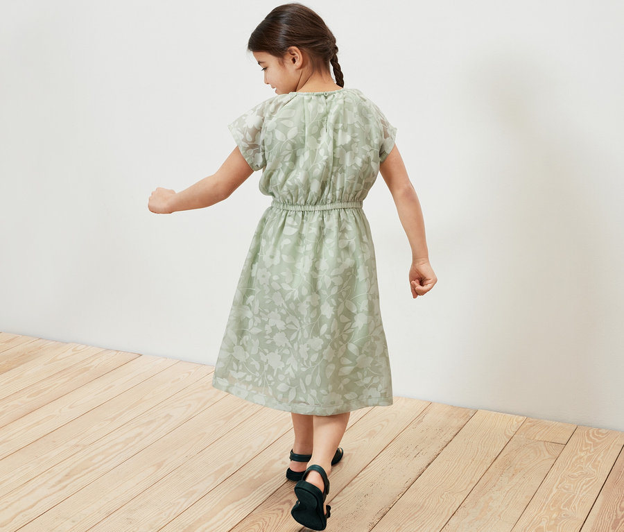 Une fille marche sur un plancher en bois, portant une robe pour enfant verte.
