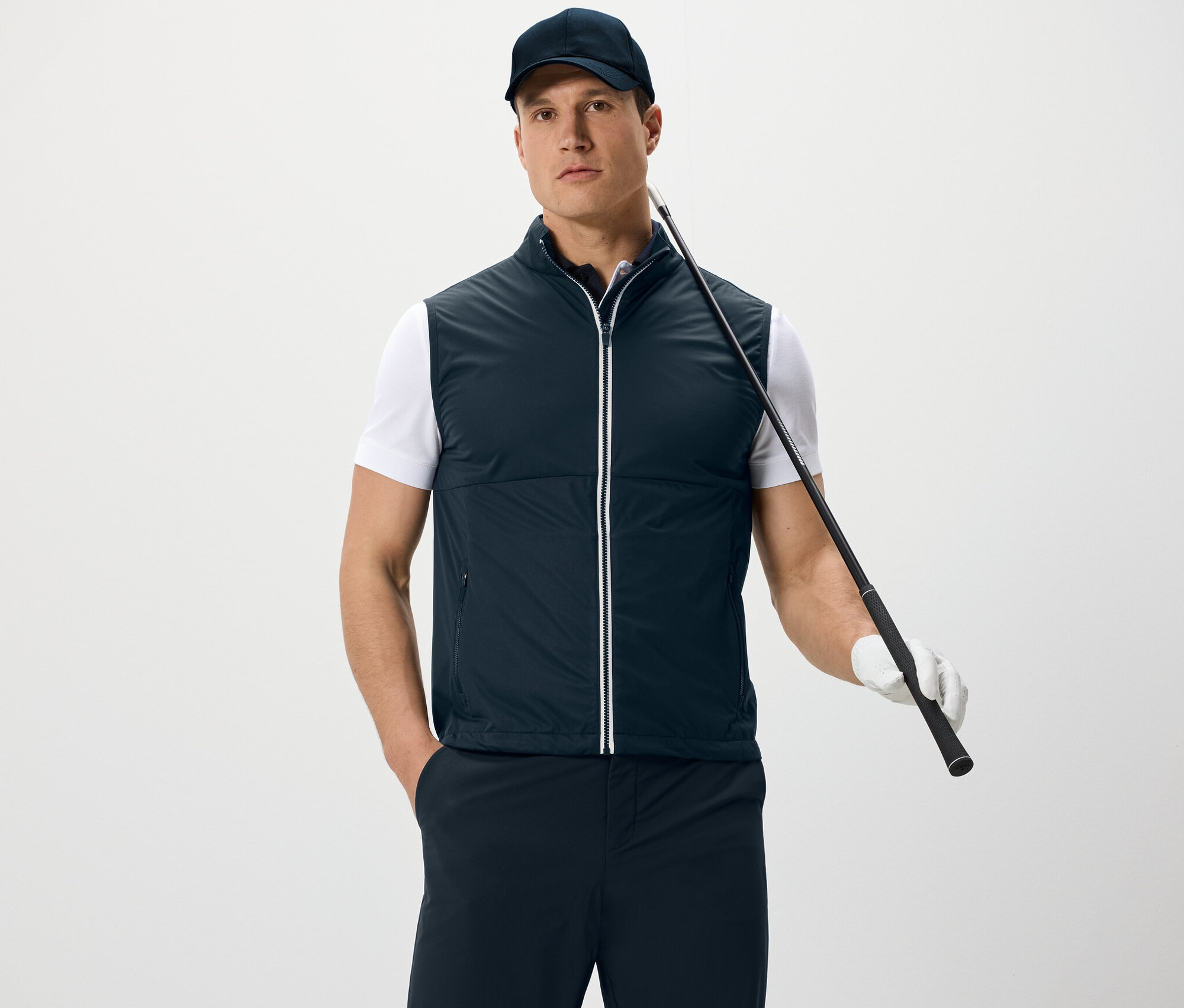 Un homme portant une veste en softshell et une casquette tient un club de golf et se tient devant un fond blanc.