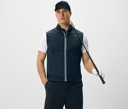 Un homme portant une veste en softshell et une casquette tient un club de golf et se tient devant un fond blanc.
