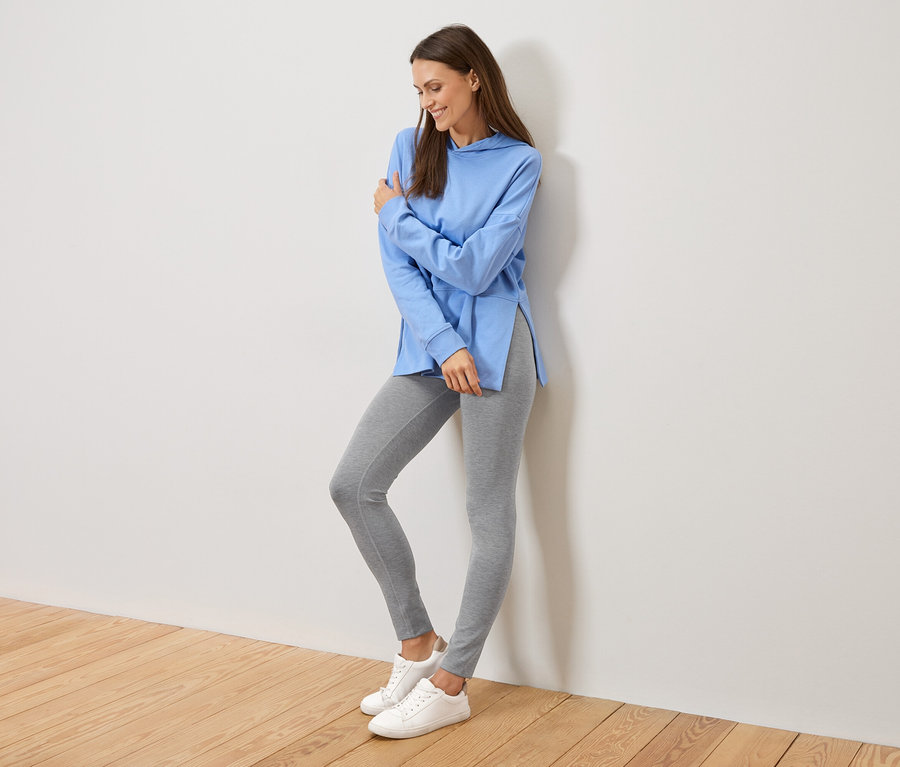 Femme appuyée contre un mur, portant un sweat à capuche bleu clair avec des fentes latérales, un legging gris et des baskets blanches.