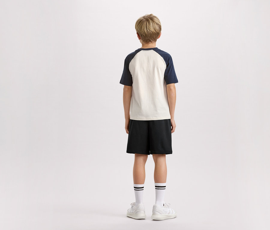 Vue arrière d'un enfant blond portant un t-shirt à manches bleu marine, un short noir, des chaussettes à rayures et des baskets blanches sur fond blanc.