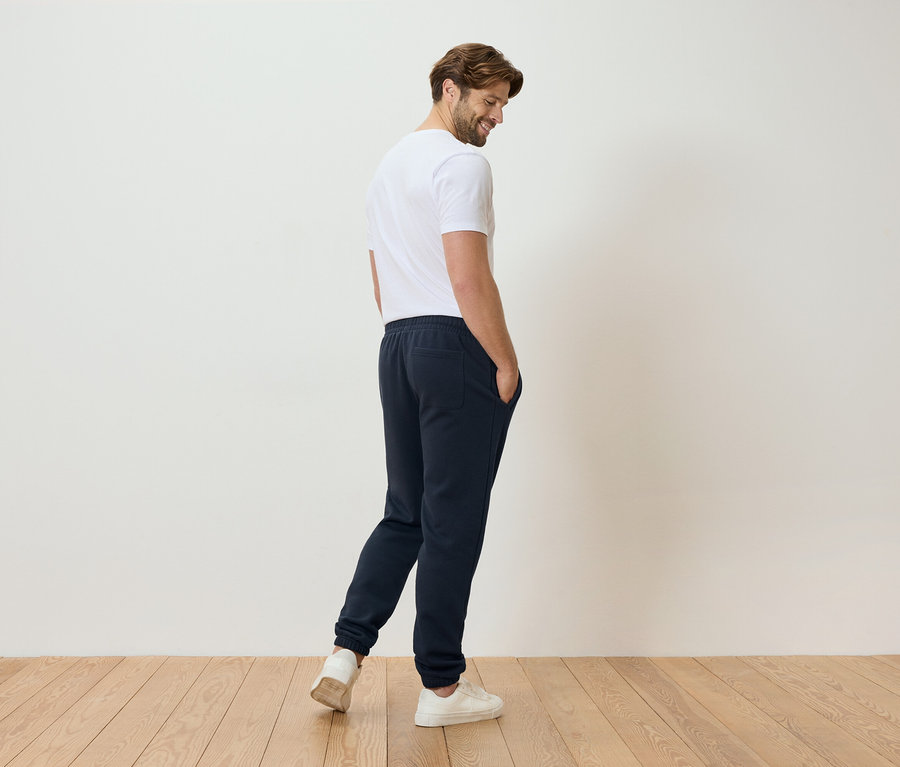 Un homme en t-shirt blanc et pantalon de jogging bleu se tient sur un plancher en bois.