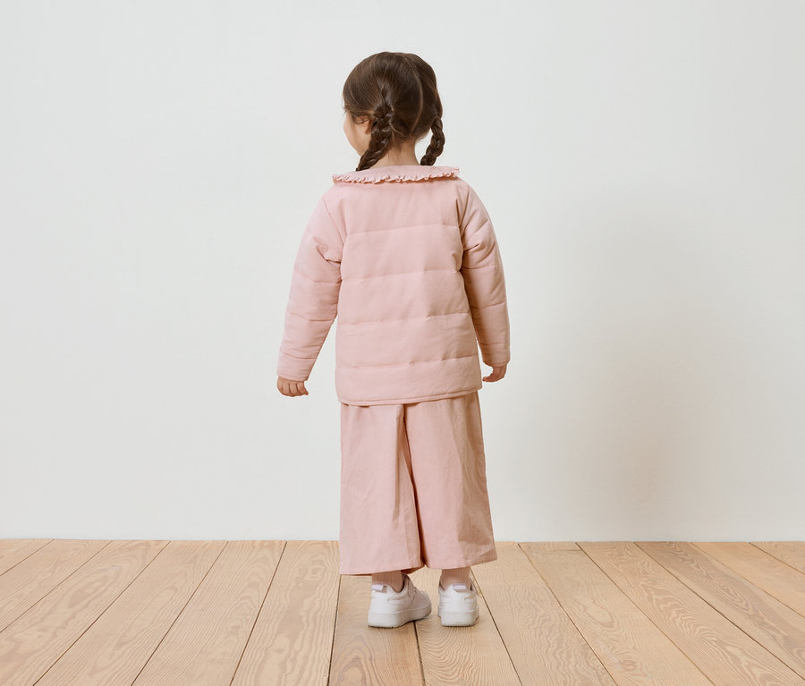 Enfant avec des tresses, vu de dos, portant une doudoune rose et un pantalon ample, avec des baskets blanches.