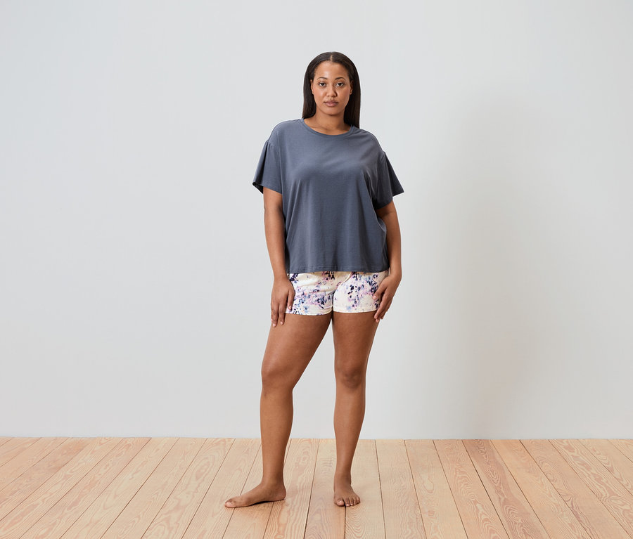 Une femme se tient pieds nus sur un plancher en bois, portant un t-shirt gris et un short à motifs floraux.