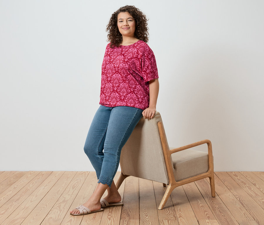 Une femme aux cheveux bouclés est assise sur une chaise, elle porte un haut rose et un jean.