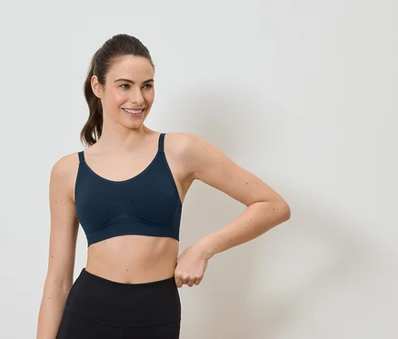Une femme souriante avec une queue de cheval porte un soutien-gorge de sport bleu marine et un legging noir sur fond blanc.