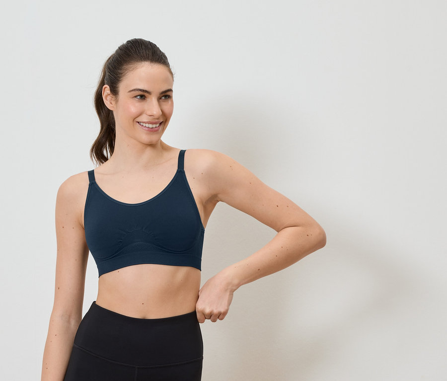 Une femme souriante avec une queue de cheval porte un soutien-gorge de sport bleu marine et un legging noir sur fond blanc.