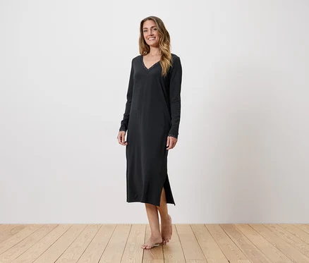 Femme portant une longue chemise de nuit sombre à col en V et manches longues, debout sur un plancher en bois devant un mur blanc.