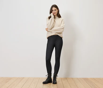 Une femme portant un pull crème, des leggings foncés et des bottes noires se tient devant un mur blanc.