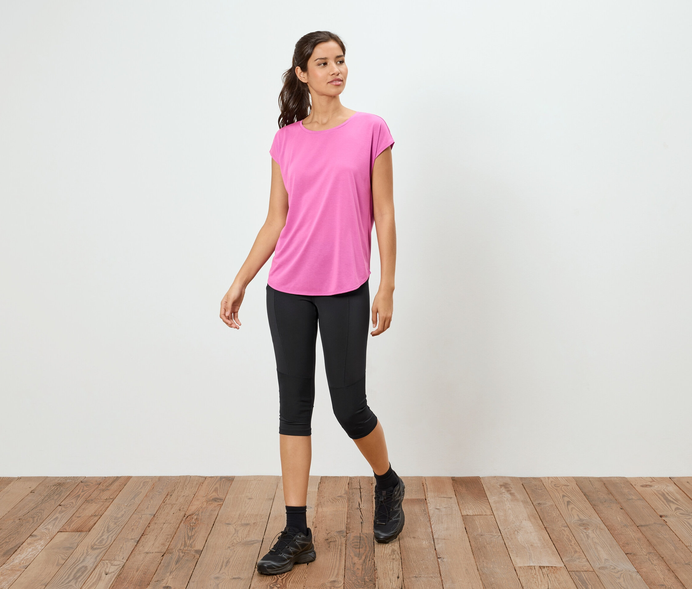 Femme debout sur un plancher en bois portant un t-shirt rose, un legging capri noir et des baskets noires.