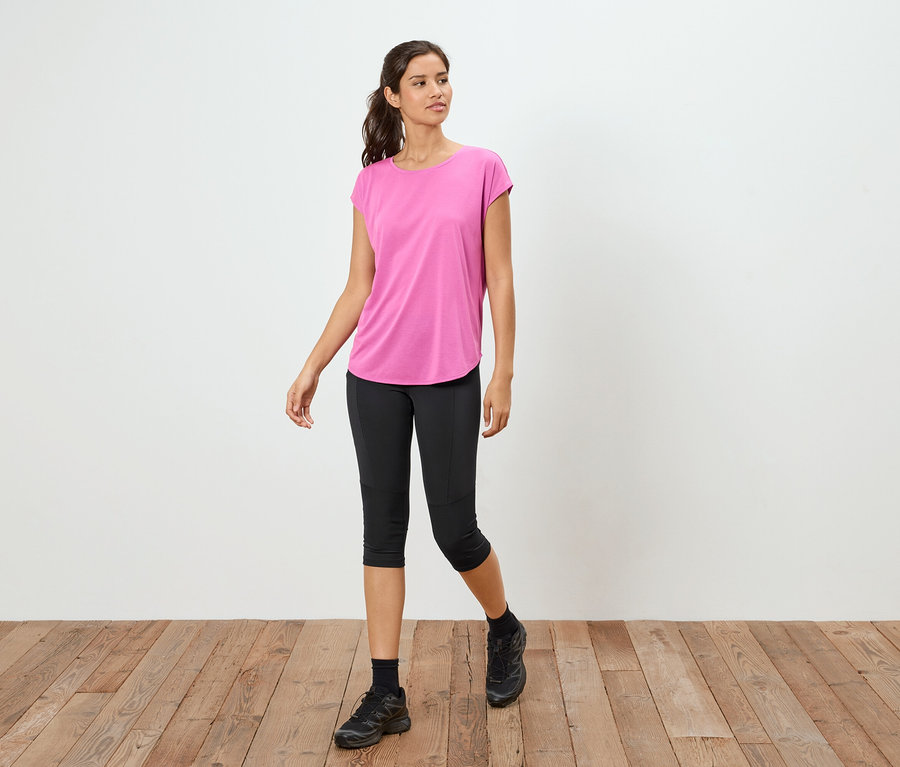 Femme debout sur un plancher en bois portant un t-shirt rose, un legging capri noir et des baskets noires.