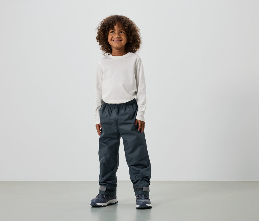 Un garçon souriant aux cheveux bouclés porte un haut à manches longues, un pantalon de pluie et des baskets.