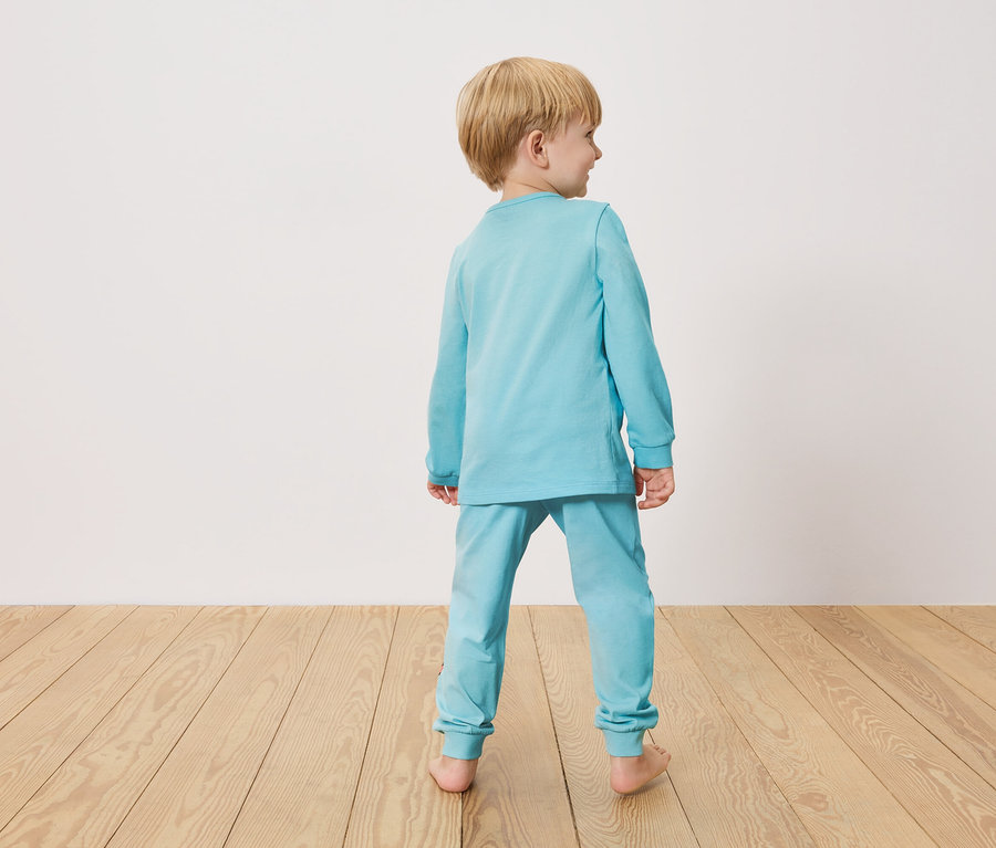 Petit garçon vu de dos, portant un pyjama bleu clair, debout sur un plancher en bois devant un mur blanc.