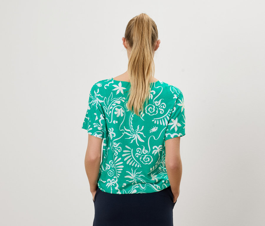 Vue de dos d'une femme blonde portant un t-shirt vert à motifs blancs et une jupe bleu marine.