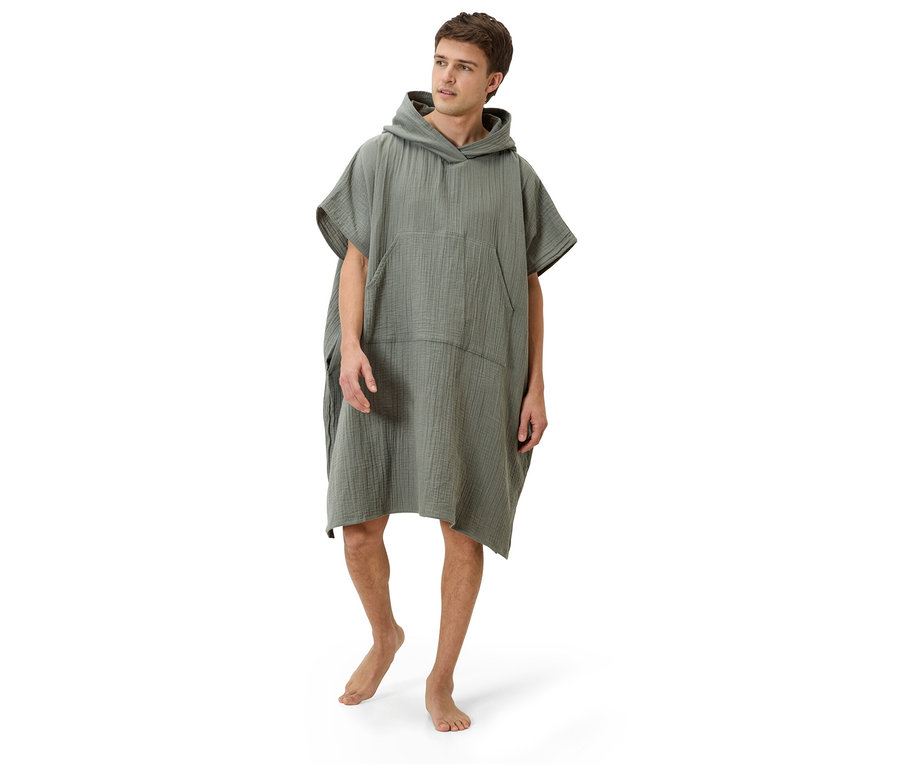 Un homme se tient pieds nus sur un fond blanc, portant un poncho à capuche vert olive.