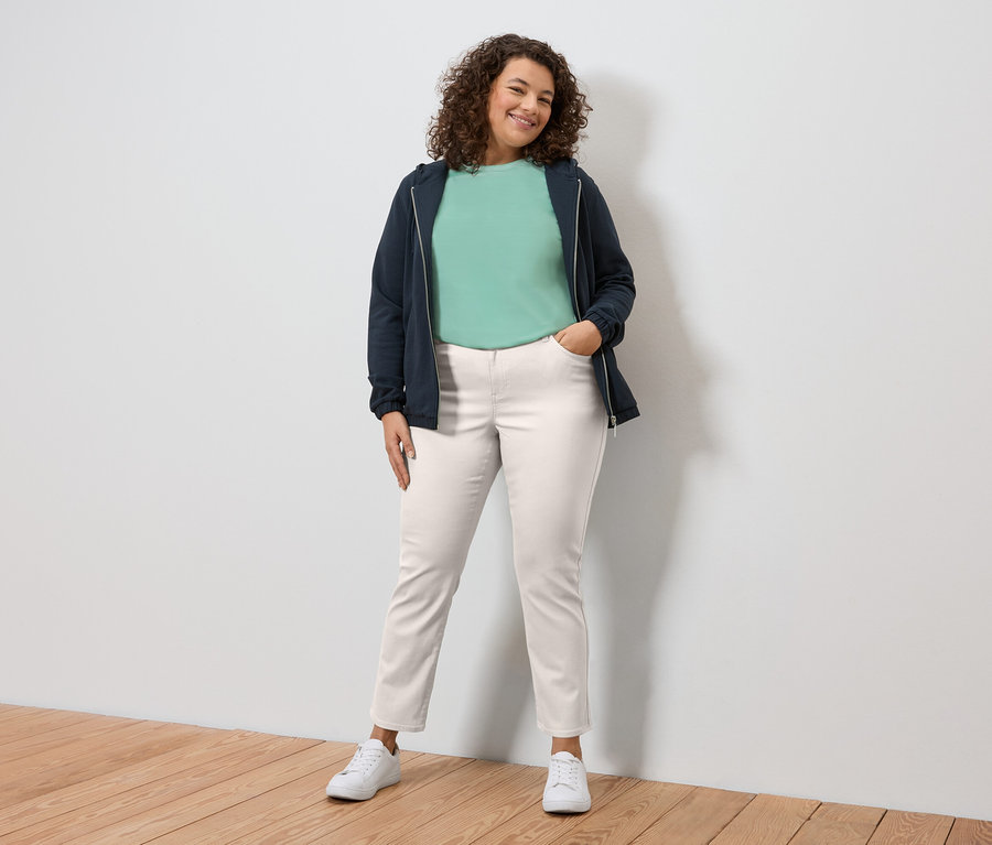 Femme aux cheveux bouclés debout devant un mur blanc, portant une veste bleu marine, un t-shirt vert, un pantalon blanc et des baskets.