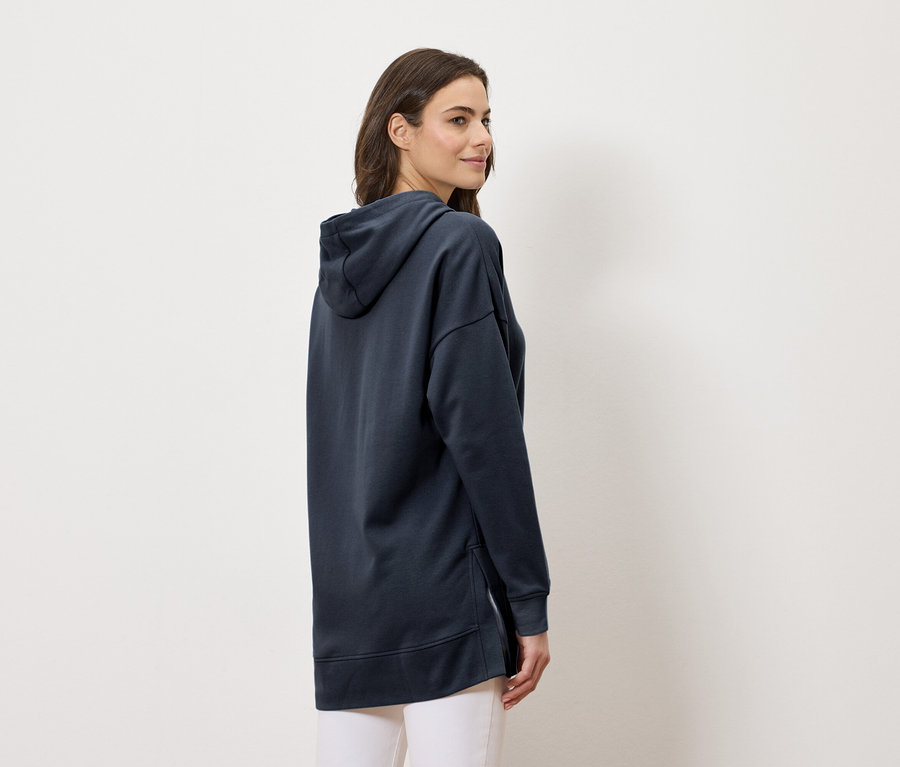 Femme portant un long sweat à capuche bleu marine et un pantalon blanc, vue de côté.