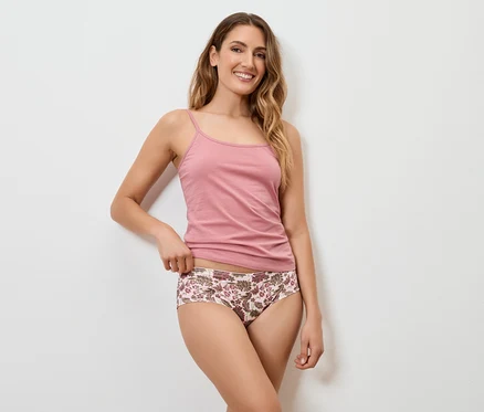 Une femme aux longs cheveux blonds ondulés sourit et pose devant un mur blanc, portant un débardeur rose et une culotte à fleurs.