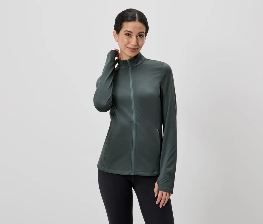 Une femme aux cheveux relevés porte une veste zippée verte et un legging noir.