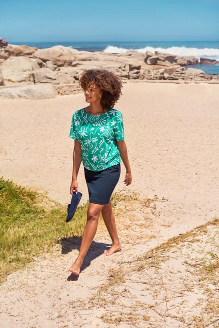 Une femme marche pieds nus sur la plage et tient des loafers en cuir velours. Elle porte des boucles d’oreilles avec pierres de cristal de verre, une jupe et un haut vert.