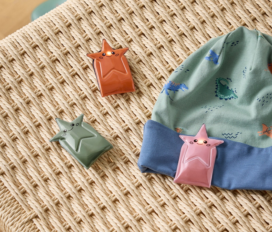 Trois clips en forme d'étoile, dont deux sont allumés, reposent sur une surface tressée beige. Un clip rose est attaché à un bonnet vert à motif de dinosaures. Un clip orange et un clip vert sont à côté du bonnet. Des bottes de pluie vertes sont en arrière-plan.
