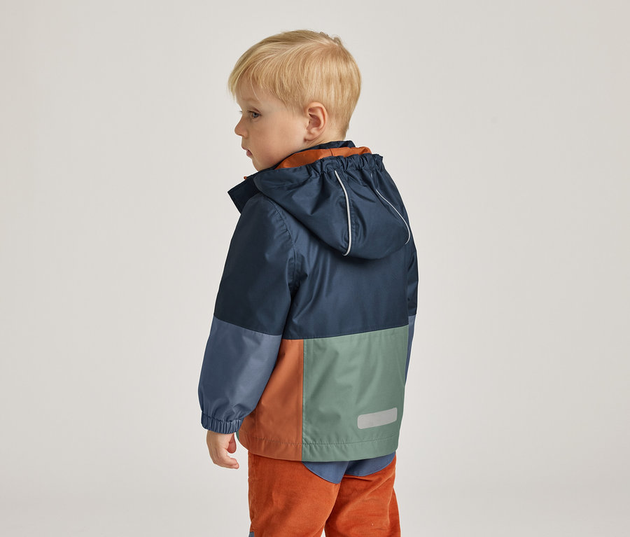 Petit garçon aux cheveux blonds courts, vu de dos, portant un imperméable à blocs de couleurs bleu, vert et orange.
