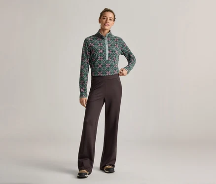 Une femme portant un haut vert à motifs et un pantalon marron pose devant un fond clair.