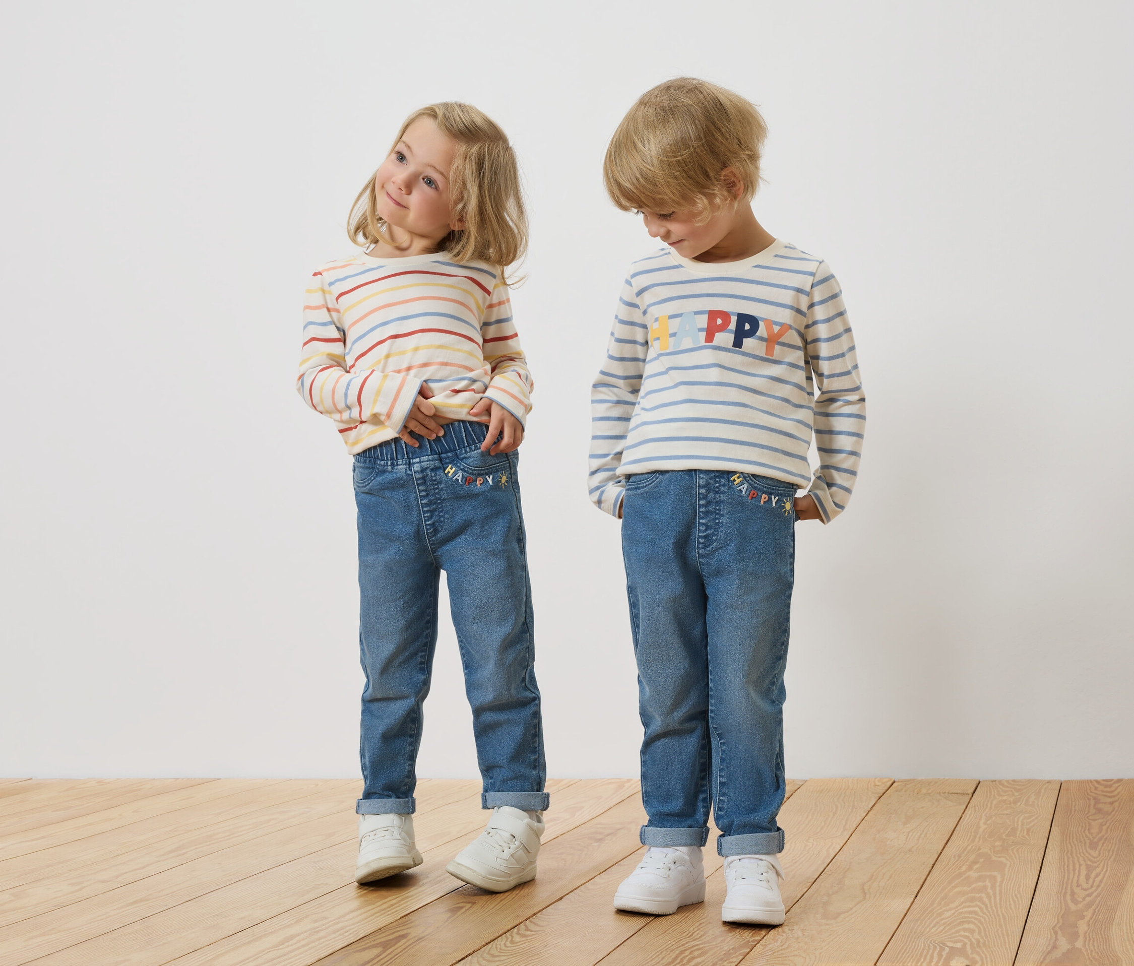 Une fille blonde et un garçon blond se tiennent côte à côte, portant des hauts à manches longues rayés et des jeans.